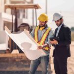 Two men in hard hats and safety glasses examining a blueprint at a construction site, discussing civil drafting plans